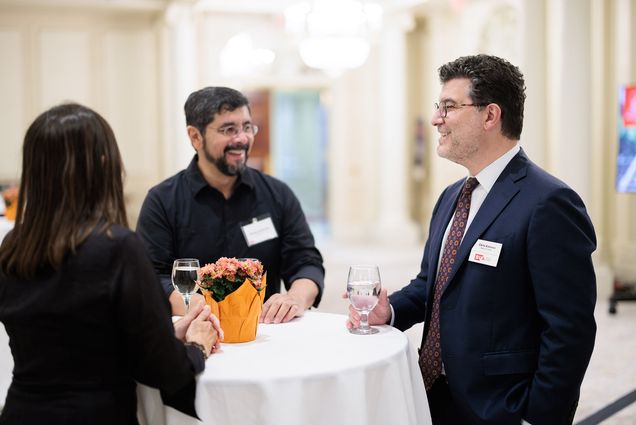Photo: Chris Kolovos, right, a white man wearing glasses, a black suit, burgundy tie and holding a glass of water, speaks to Susanna Lara, a woman with brown hair and black blazr facing away from the camera, left, and Horacio Gutierrez, A Hispanic man wearing glasses and black long-sleeved collared shirt, middle. They all chat and stand around a white high-top table.