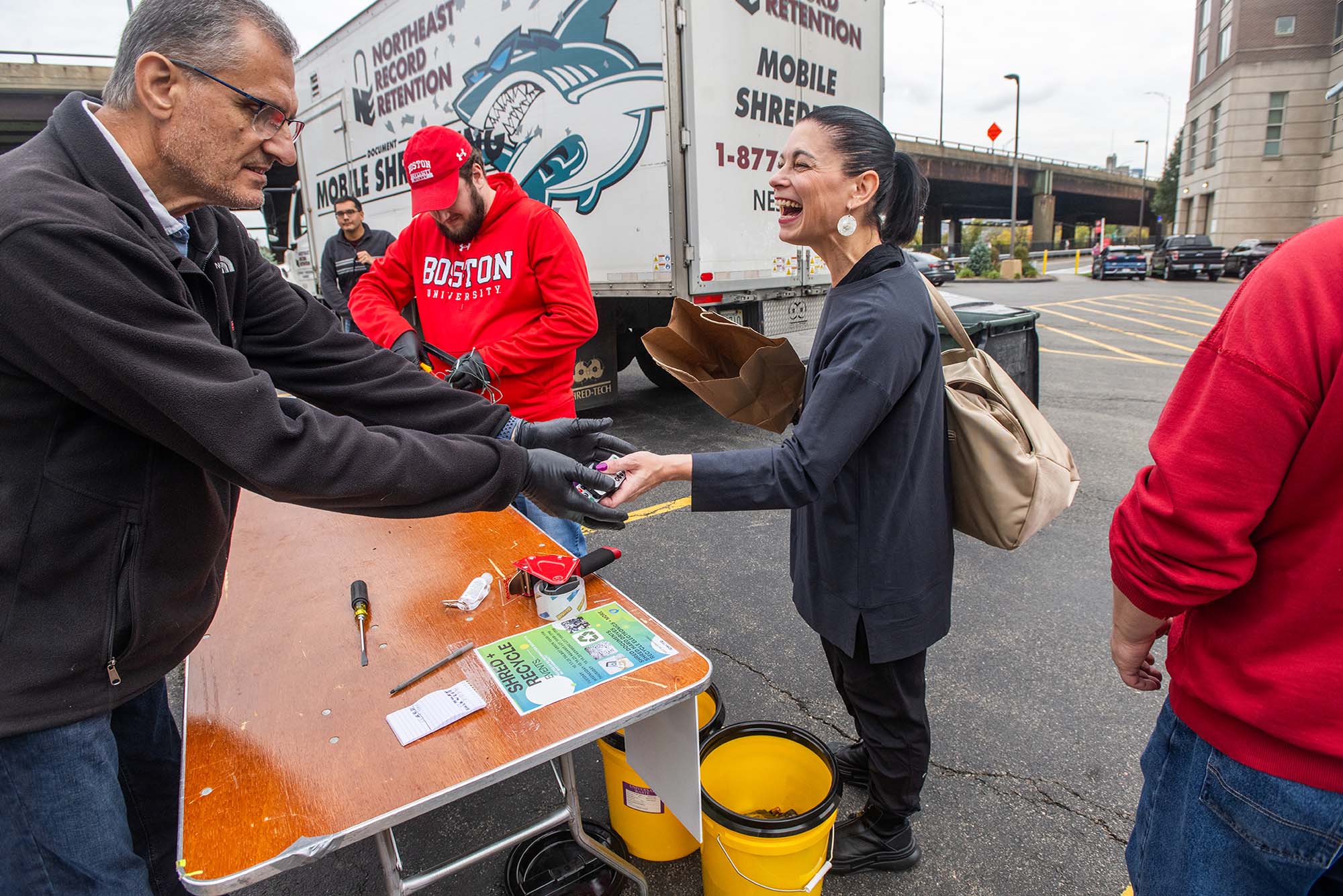 Photo: A woman hands a bag of batteries to a man wh stands behind a wooden table set up in a vacant parking lot. A large white truck can be seen behind them.