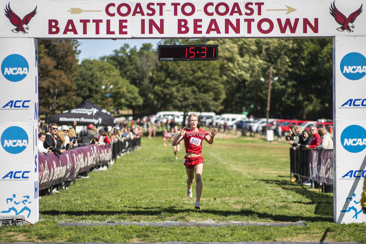 Photo: Aksel Laudon, a white young man wearing a red BU cross country uniform, raises his arms as he runs across the finish line on the Battle in Beantown race on a sunny day.