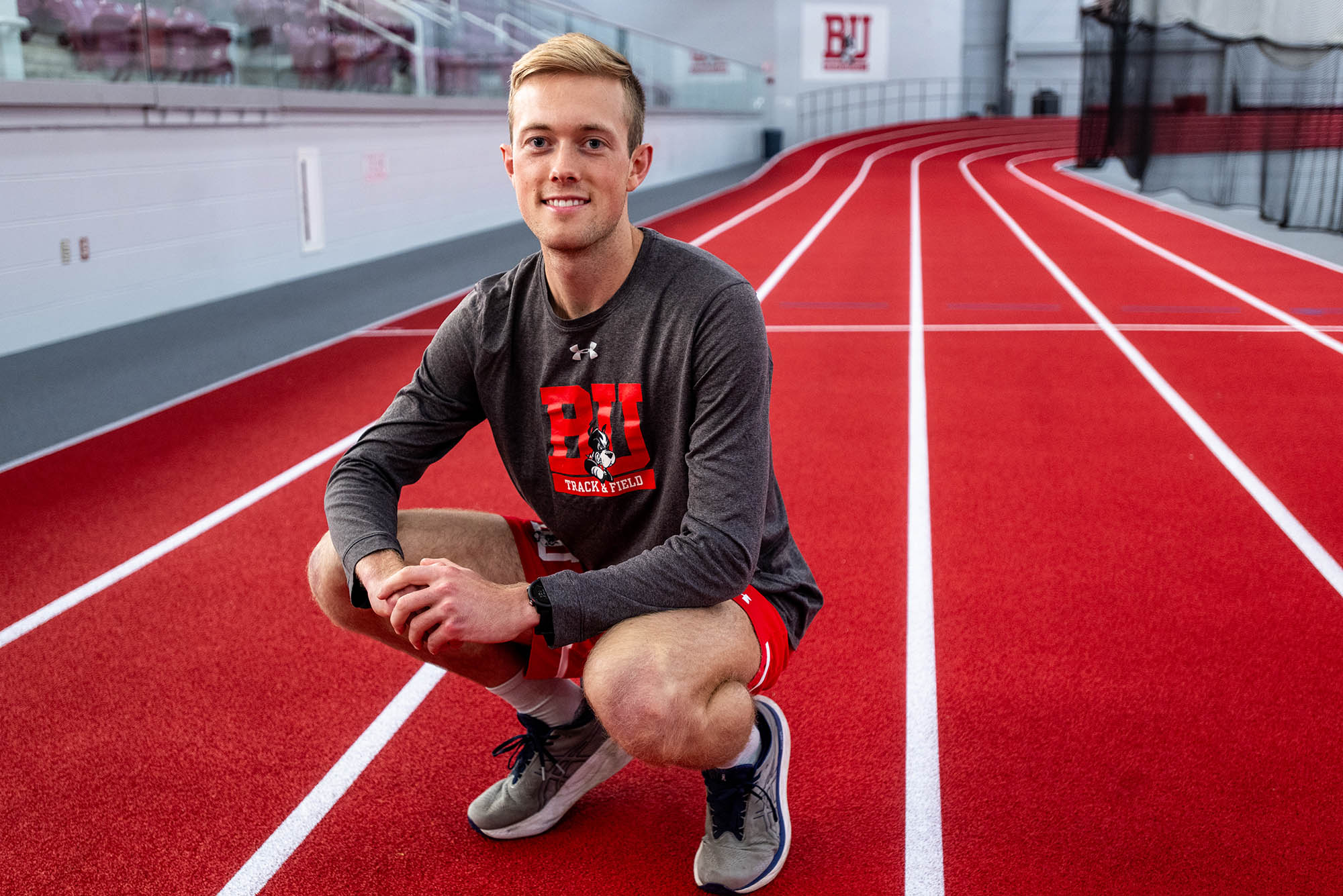 Photo: Aksel Laudon, a young white man with blonde-brown hair and wearing a grey long-sleeved BU XCTF shirt and red shorts squats on the red indoor track lanes of the BU Track and Tennis center.