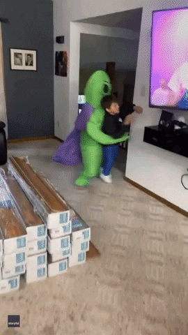 This is a GIF that is taken within a person's home a little boy with an alien costume on. The alien is green and blue on the button and is blown up on the back of the little boy that is wearing it.He is wearing a black shirt and is running behind the wall to hide from the camera. He accidentally walks into the wall and falls for a second and gets back up and runs behind the other wall.