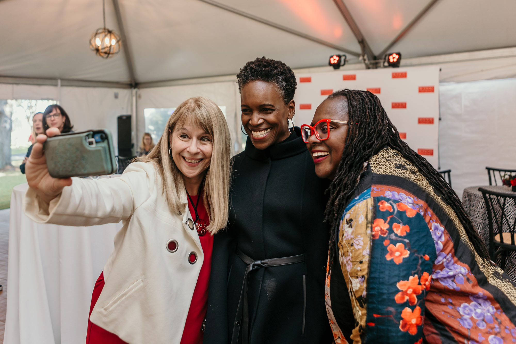 Photo: Barbara Jones, a white woman with blonde hair and wearing a red blouse and white blazer (left), and Angela Onwuachi-Willig, a Black woman with long dreadlocks wearing red glasses and a colorful, patterned long-sleeved top, (right), pose for a selfie with Melissa Gilliam (center), a Black woman wearing a black jacket and pants.