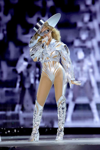 This is a photo of the famous Beyonce while she is performing in concert. She is standing on stage in front of a grey, blurred background. She is wearing a fully metallic outfit. She has on a metallic hat that is sideways on her head, futuristic silver sunglasses, a stringy silver body suiit that has sleeves, and metallic boots that connect to her body suit through a strap that comes up the sides of her legs. She has one hand by her side with her palm facing the floor while her other hand grips the microphone to her mouth.