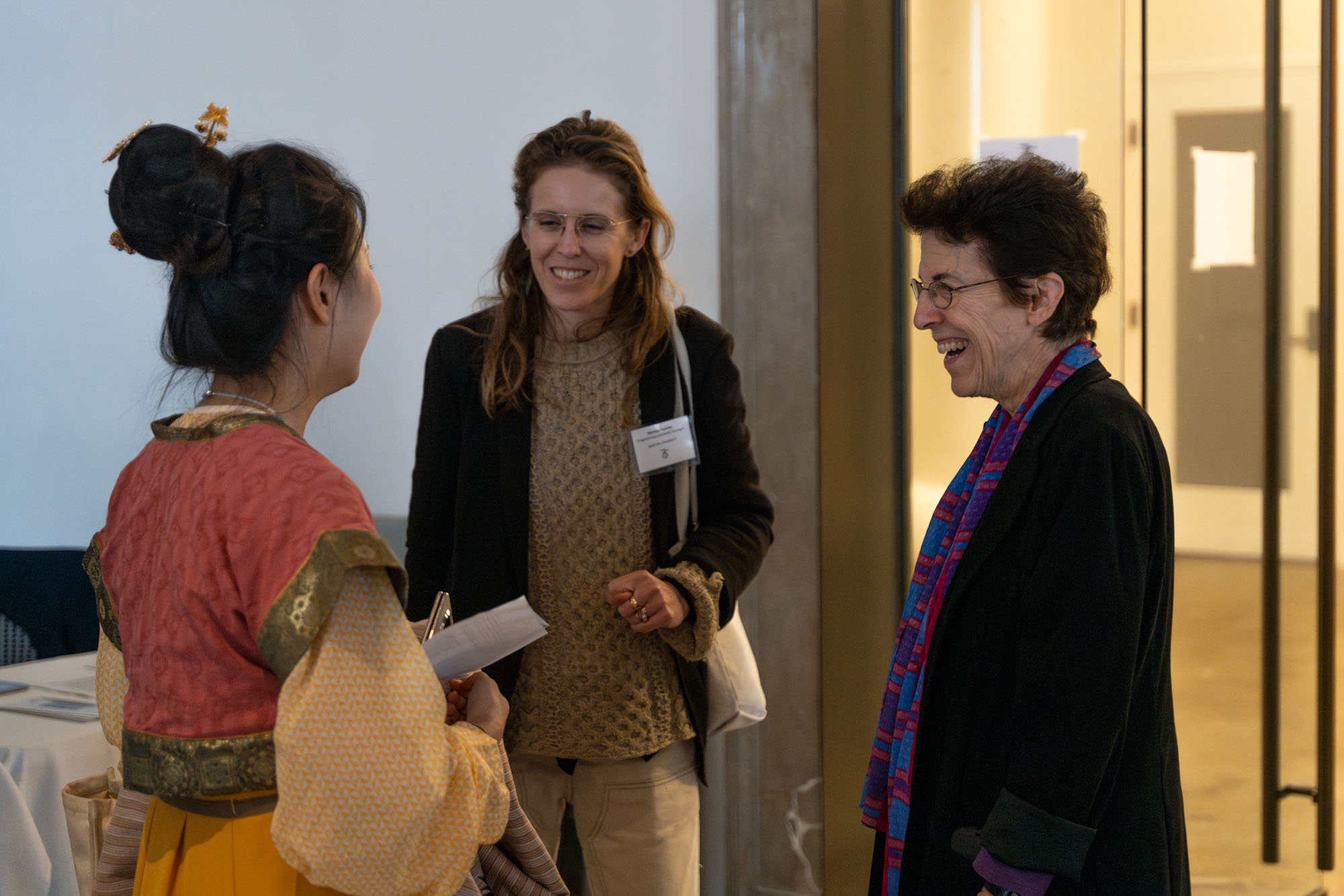 Photo: Deborah Cornell (right), a white woman with short brown hair and wearing glasses, a black blazer, and purple scarf, smiles and chats with two patrons to her left in a gallery.
