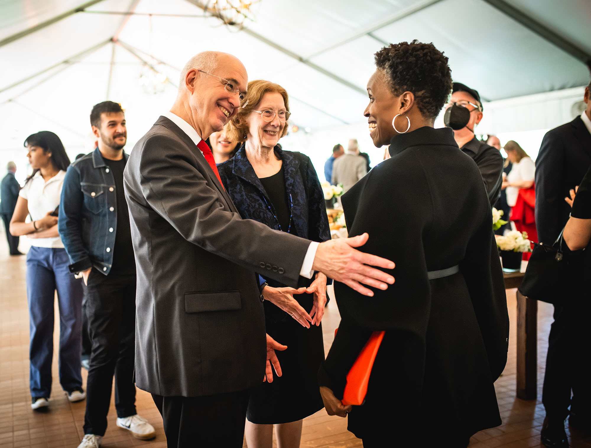 Photo: The new BU president, Melissa L. Gilliam (right), a Black woman wearing a black jacket and pants, greets Interim president Ken Freeman (left), a tall white man wearing a black suit and red tie, and his wife, Janice (center), a white woman wearing a black dress and blazer, during a reception.