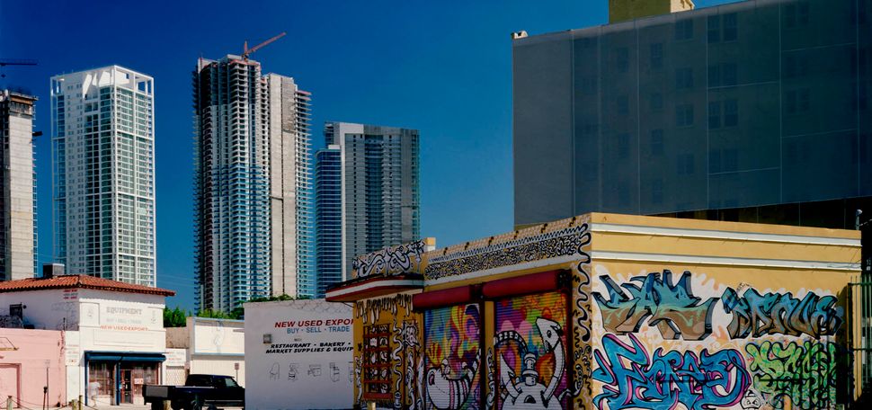 Photo: an urban downtown scene of the Miami cityscape is shown. A small building covered in graffiti is juxtaposed in the foreground against new, tall glass buildings behind it.