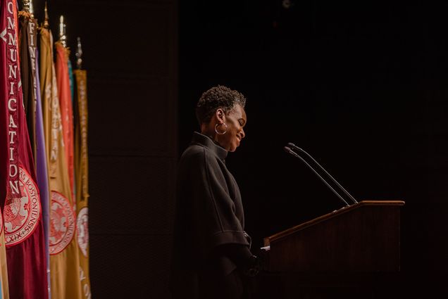 Photo: Dr. Melissa L.Gilliam, a Black woman with a short afro and wearing a black turtleneck sweater, speaks into a microphone on a wooden podium. Sign on podium reads "Boston University". Behind her, a row of colorful flags are placed.