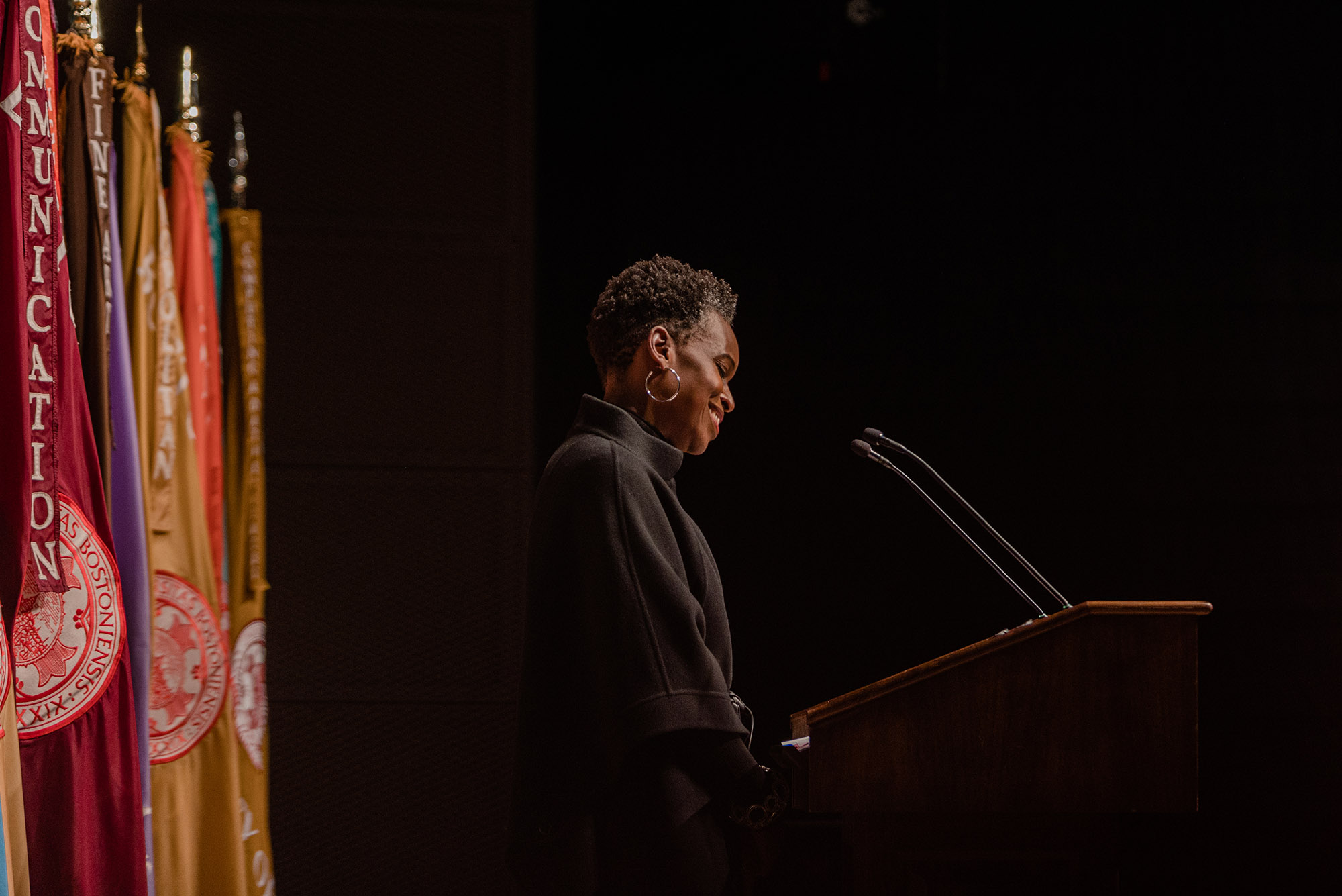 Photo: Dr. Melissa L.Gilliam, a Black woman with a short afro and wearing a black turtleneck sweater, speaks into a microphone on a wooden podium. Sign on podium reads "Boston University". Behind her, a row of colorful flags are placed.