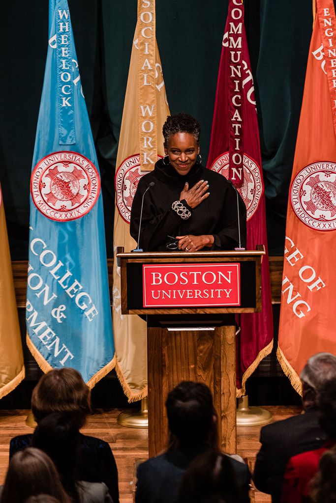 Photo: Dr. Melissa L. Gilliam, a Black woman with a short afro and wearing a black turtleneck sweater, speaks into a microphone on a wooden podium. She smiles and holds a hand over her heart as she looks down to the audience. Sign on podium reads "Boston University". Behind her, a row of colorful flags are placed.