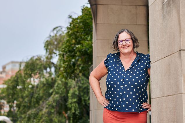 Photo: A woman with glasses and a polkadot blouse and red skirt poses for a picture in front of a stone building on a sunny day