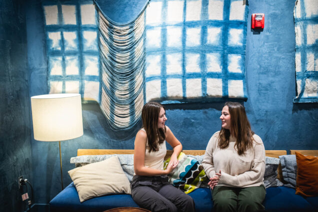 Photo: Two women sit on a couch, facing each other while talking. The wall behind them is a vivid blue with a fabric art piece with a checkered pattern. In the middle of the piece, the fabric is frayed to create a draping effect.