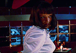 This is a GIF of character Mia Wallace from Pulp Fiction. She appears to be in a public place that may be a bar/restaurant that has dim lighting. She has short black hair that is cut into a bob with bangs. She is thin and is wearing a white shirt, a black bra that is slightly exposed, and is wearing red lipstick. She is intensely staring at the person in front of her while she sways her shoulders back and forth.