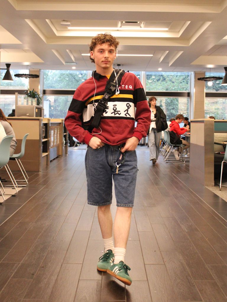 Photo: A student in a red shirt and grey shorts stands with hands in pockets