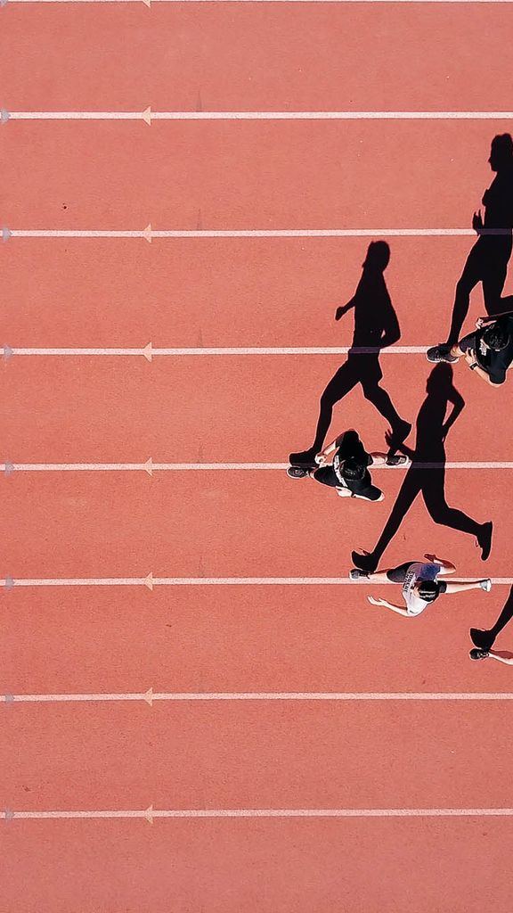 Photo: A group of runners running in a arrow formation on a outdoor track. The photo is from a birds eye view.
