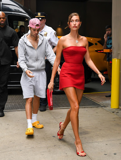 This is a photo of Hailey and Justin Bieber that was taken by paparazzi. The couple is walking into Haileys brand event for the launch of her new product. They are on a city sidewalk, with security and the car they got out of behind them. Hailey is walking in front of Justin, with her light brown hair in a high bun, a red strapless mini dress on, red kitten heels, with a red leather purse to match. She is looking out in the distance with a straight face. Justin is behind her in a grey suit suit, wearing a zip up grey hoodie zipped half-down exposing his tattoos and light grey shorts on. He has medium length white socks on, with bright yellow Crocs on his feet. He is wearing a light pink hat that is backwards. He is looking down in front of himself.
