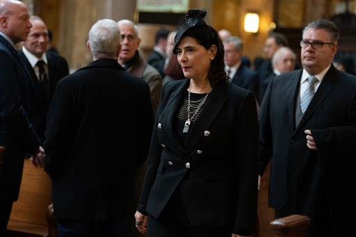 This photo is from the show Succession that aired on HBOMAX. There is a group of people all wearing black and talking to one another. In front of the group, there is a woman standing alone, looking off into the distance. She has black hair, dark eyes, and lipstick on. She is wearing all black; she has a black dress on, a black pea coat, and a black fascinator on. 