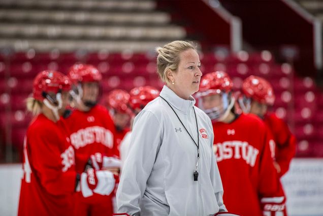 Photo: A blonde woman wearing a whistle around her neck stands in front of a group of womens hockey players in red jerseys