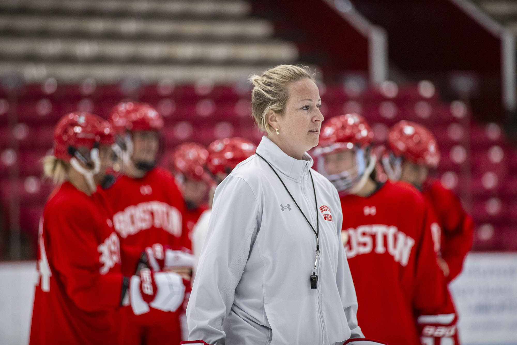Photo: A blonde woman wearing a whistle around her neck stands in front of a group of womens hockey players in red jerseys