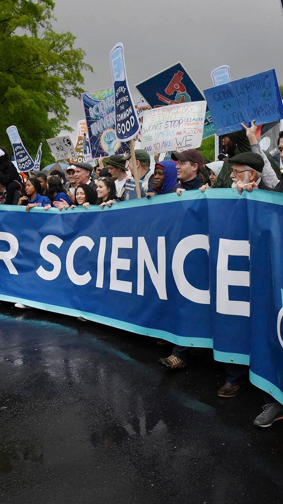 Photo: Thousands of scientists and their supporters join the March for Science in Washington, D.C. A crowd of people hold signs and chant on a rainy, gloomy day. A row of people int he front hold a large blue banner across them that reads "March for Science".