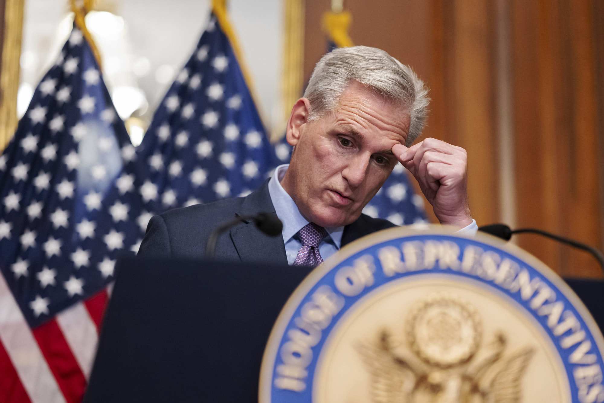 Photo: Kevin McCarthy, former House Speaker, rubs his forehead with his thumb as he looks down, dejectedly. He is a white man with a black suit, blue button up, and a red and blue tie. He stands at a podium with the United States emblem on it and the American flag in the background.