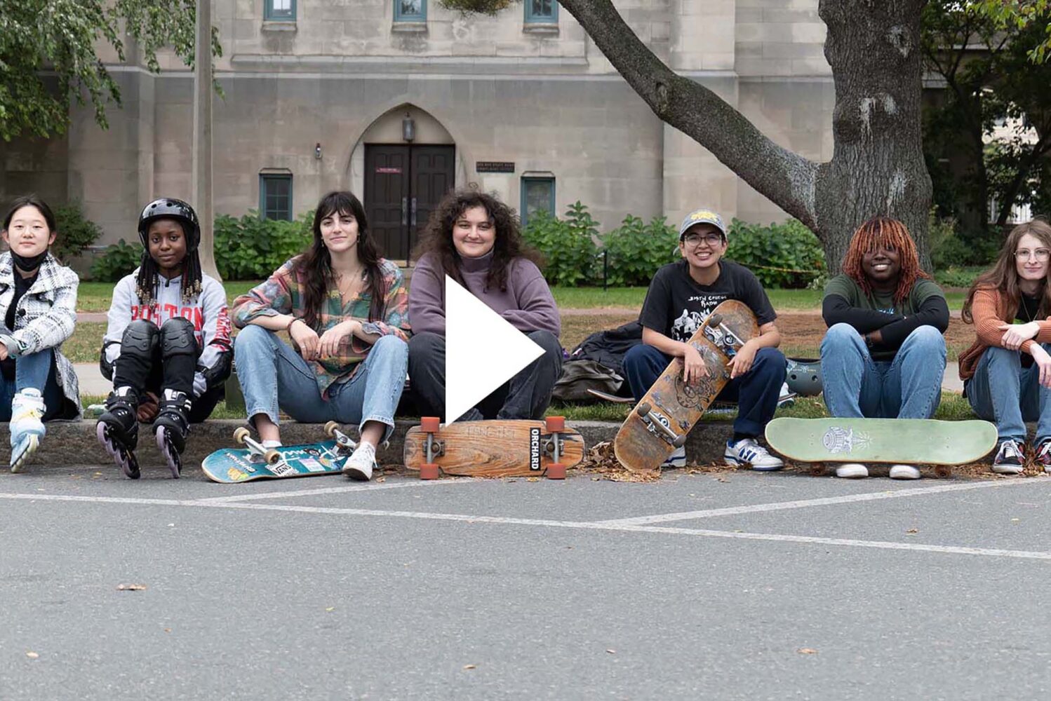 Photo: The BU Girls' Skateboarding Club poses for a photo along the BU Beach, an outdoor area where BU students can relax and unwind outside. A video play button overlay rests on top of the photo.