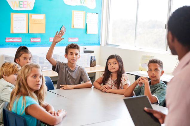 Photo: A table of children sit in a circle. Some have their arms on the table, some don't. A boy, in the center of the photo with a gray shirt, raises his hand.