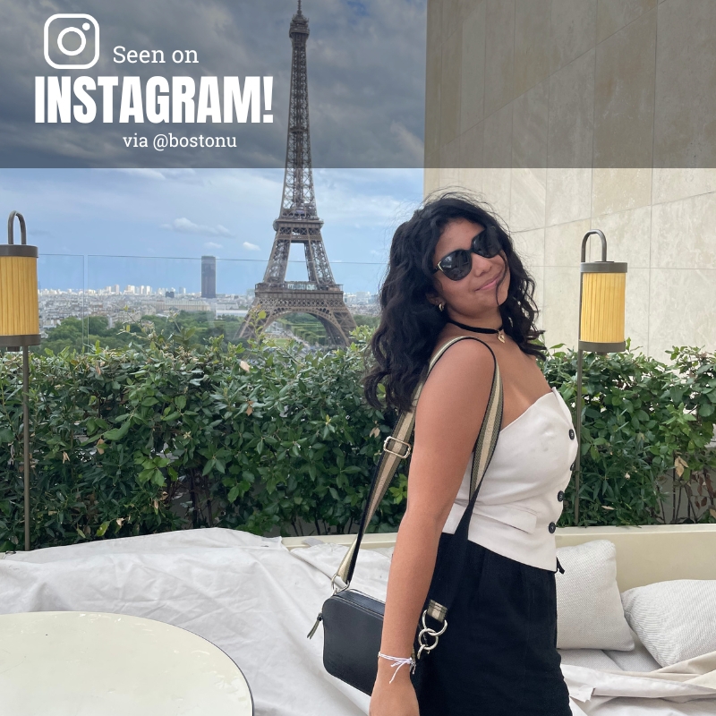 Photo: A young woman poses on a balcony that has a perfect view of the Eiffel Tower. She wears a chic white top, black pants and sunglasses, and smiles for the camera.