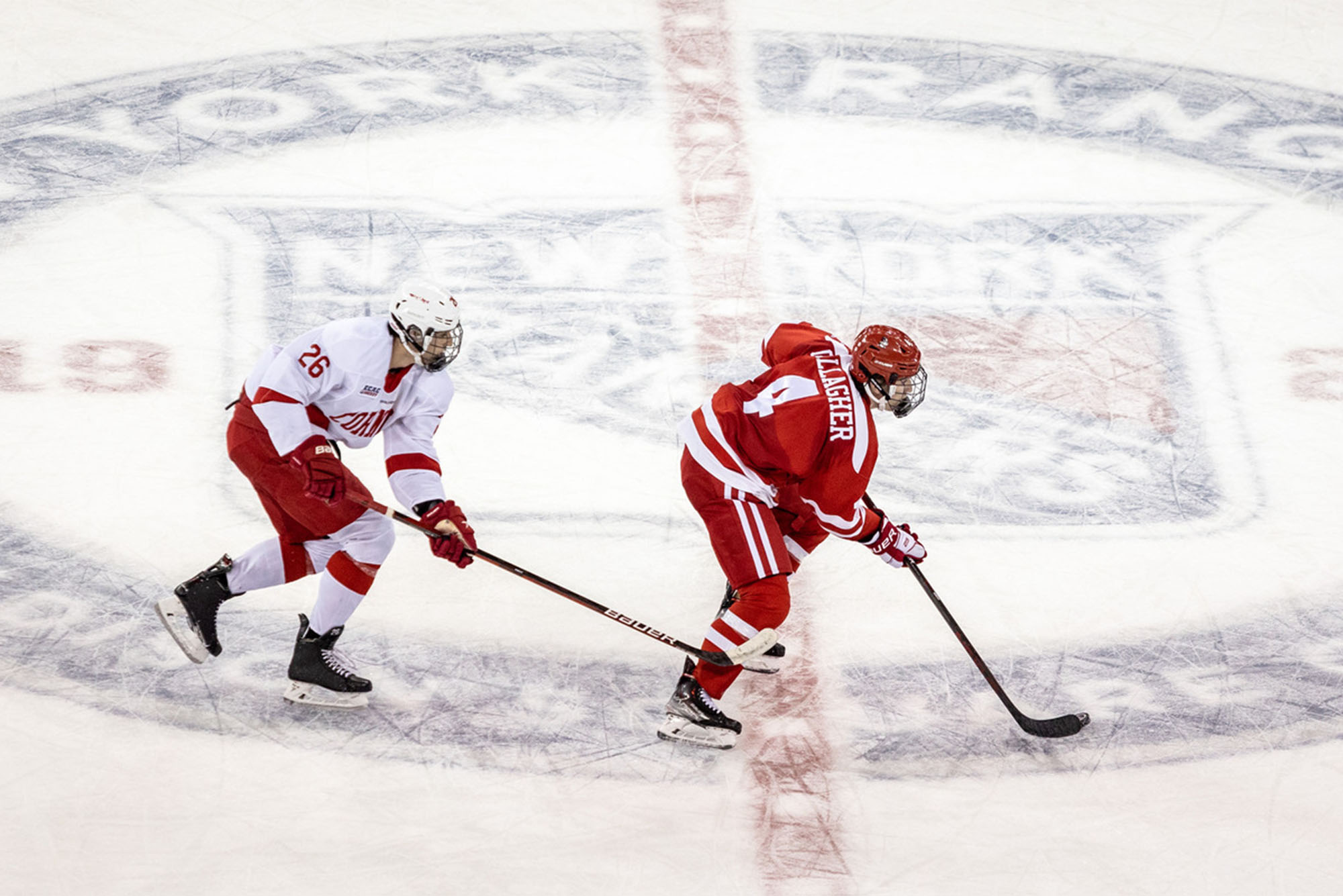 Photo: Two hockey players chase each other, one has the puck in a red uniform and the other is wearing white. They are near center ice at Madison Square Garden.
