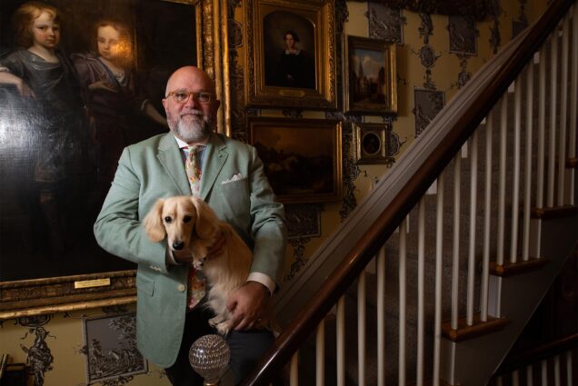 Photo: A bald man in a suit holds a dachshund while standing on an ornate wooden staircase in front of many old time portrait paintings