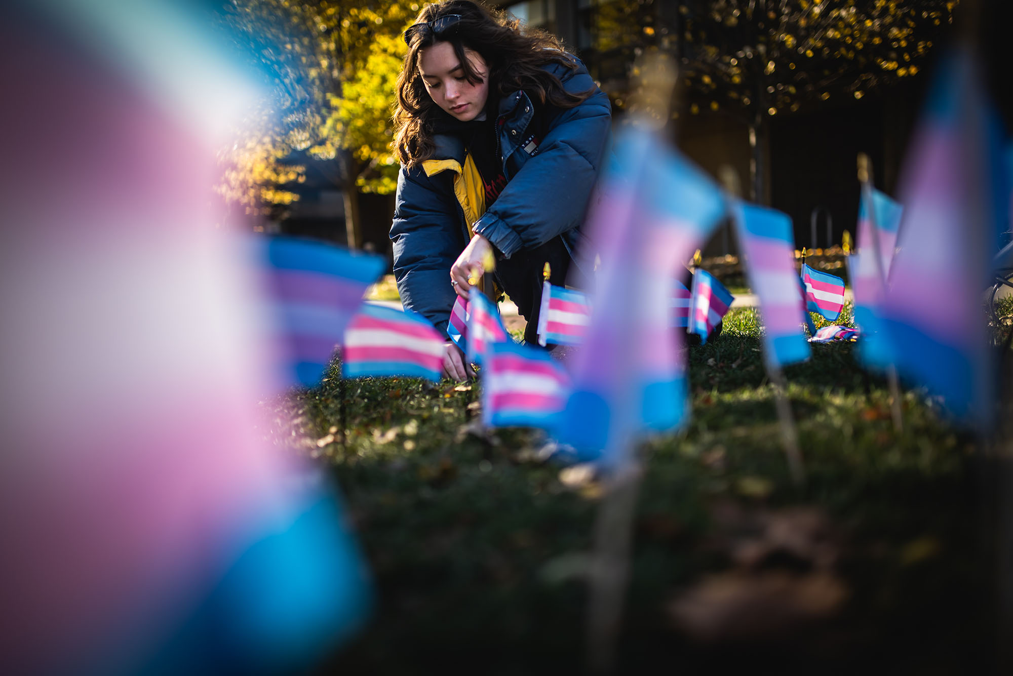 Photo: A young woman wearing a puffy, dark blue jacket places a small light, pink, and white striped flag into the ground. Around her are similar Transgender flags placed in the ground as well.
