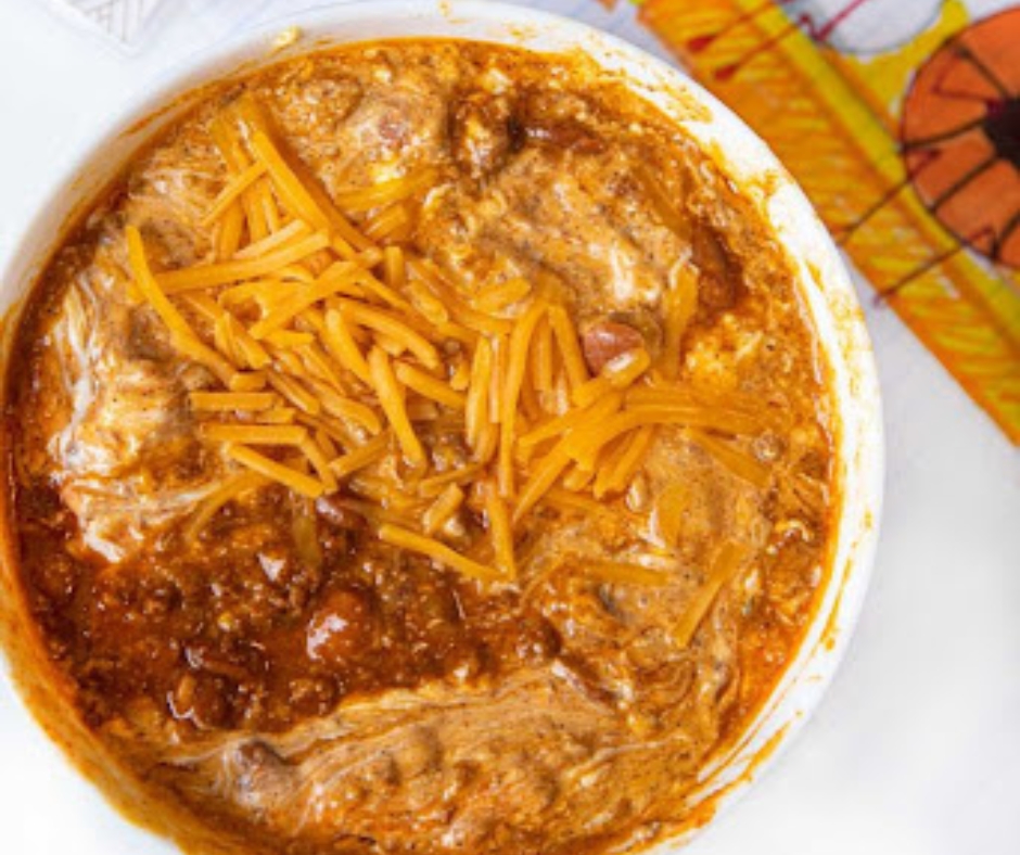 This is a cheesy chili dip. It is in a white bowl and has red salsa and shredded orange cheese.