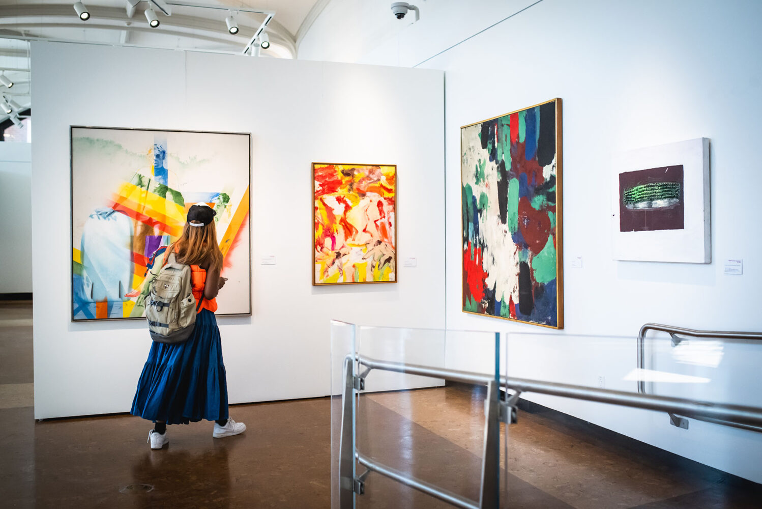 Photo: A large, white gallery wall features large paintings from various artists. A person is showing walking through and looking at different pieces.