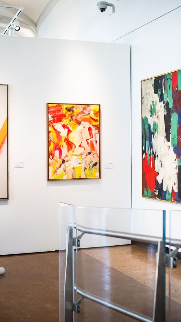 Photo: A large, white gallery wall features large paintings from various artists. A person is showing walking through and looking at different pieces.