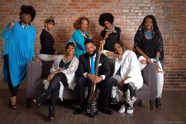 Photo: A group of eight Black people, three sitting on a counch in the center of the room and five standing eahind and to the sides of the couch, pose and smile for the photo. They all wear clothes that are black, white, or blue and are featured in front of a brick wall. The person in the center holds an electric guitar in their hands.