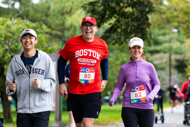 Photo: Three people wearing are shown as they run during a 5k. From left to right: A younger Asian man wearing a grey sweatshirt, an older white man wearing red Boston University shirt and cap, and a woman wearing a purple jacket run towards the camera.