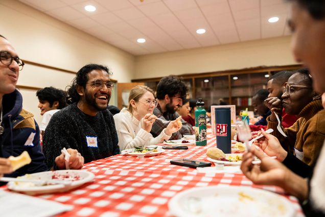 Photo: A large group of diverse people sit at a red and white checkered table as they laugh, chat, and eat a Thanksgiving meal.