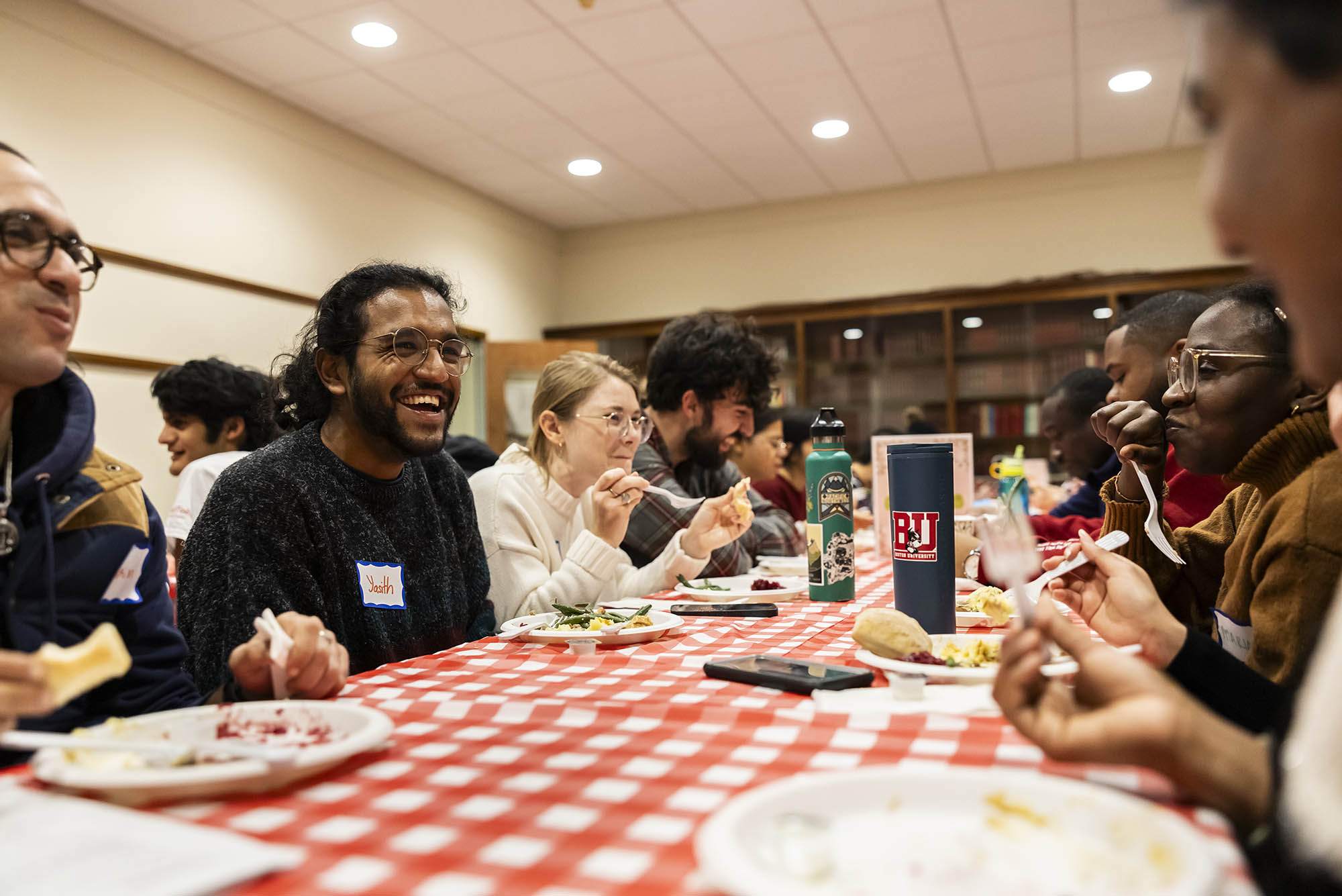 Photo: A large group of diverse people sit at a red and white checkered table as they laugh, chat, and eat a Thanksgiving meal.