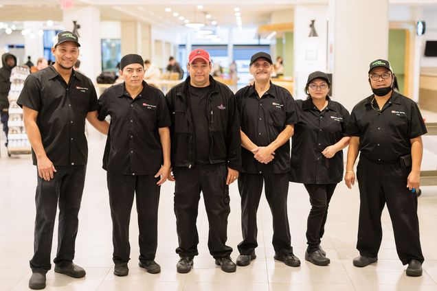 Photo: Six people dressed in black Boston University dining worker uniforms smile and pose together. From left: Carlin Ventura, Ilder Montesanos, Luis Morales, Ramiro Contreras, Bertilla Reyes, and Jose Amador stand for a photo after a brief team celebration. They stand in a brightly lit area of a dining hall.