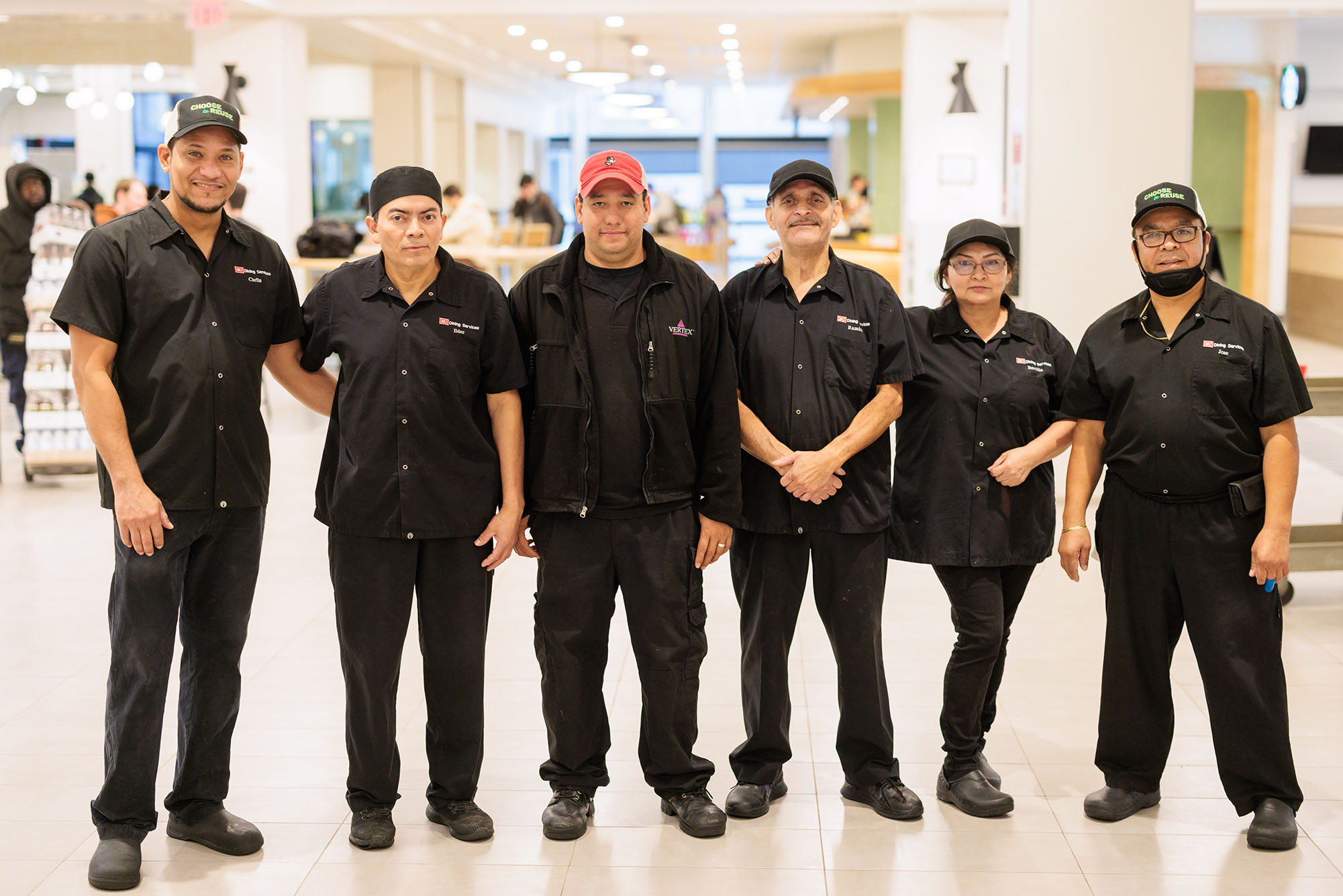 Photo: Six people dressed in black Boston University dining worker uniforms smile and pose together. From left: Carlin Ventura, Ilder Montesanos, Luis Morales, Ramiro Contreras, Bertilla Reyes, and Jose Amador stand for a photo after a brief team celebration. They stand in a brightly lit area of a dining hall.