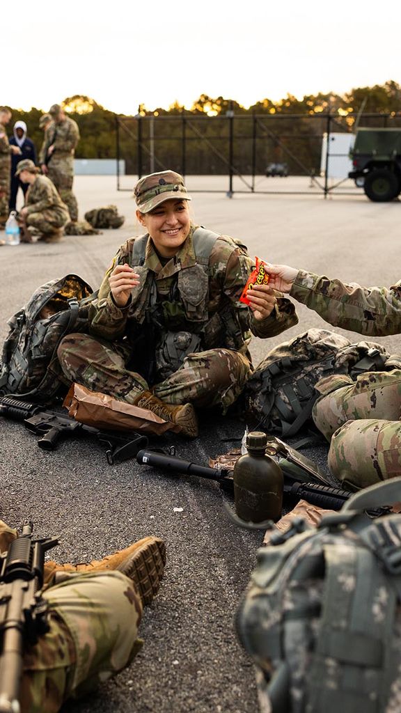 Photo: ROTC Army Cadet, Audrey Soukup (Sargent’25) (second from right) and Dharma Cooper (Pardee’25) share Reeces Pieces candy while they wait with their platoon. A small group of young women dressed in army ROTC gear sit on the ground and share candy with each other.