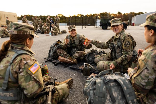 Photo: ROTC Army Cadet, Audrey Soukup (Sargent’25) (second from right) and Dharma Cooper (Pardee’25) share Reeces Pieces candy while they wait with their platoon. A small group of young women dressed in army ROTC gear sit on the ground and share candy with each other.
