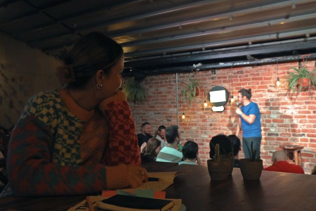 Photo: In the foreground, a young woman wearing a colorful sweater and taking notes in a notebook looks to her left towards a comedian on stage, telling jokes under a spotlight to a small audience in front of them.