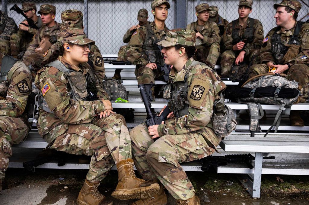 Photo: ROTC Army Cadet Dharma Cooper (left) and Audrey Soukup chat while waiting. Two young women wearing Army ROTC gear sit on metal benches with other cadets wearing the same uniform. They face each other and chat as they sit.