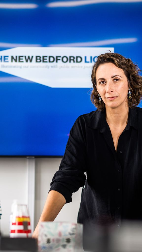 Photo: Daniela Melo, a white woman with short, curly brown hair and wearing a black, long-sleeved collared shirt leans forward against a white counter with various lighthouse and editorial paraphernalia on it. Behind her a large tv screen displays the blue lighthouse logo of "The New Bedford Light"