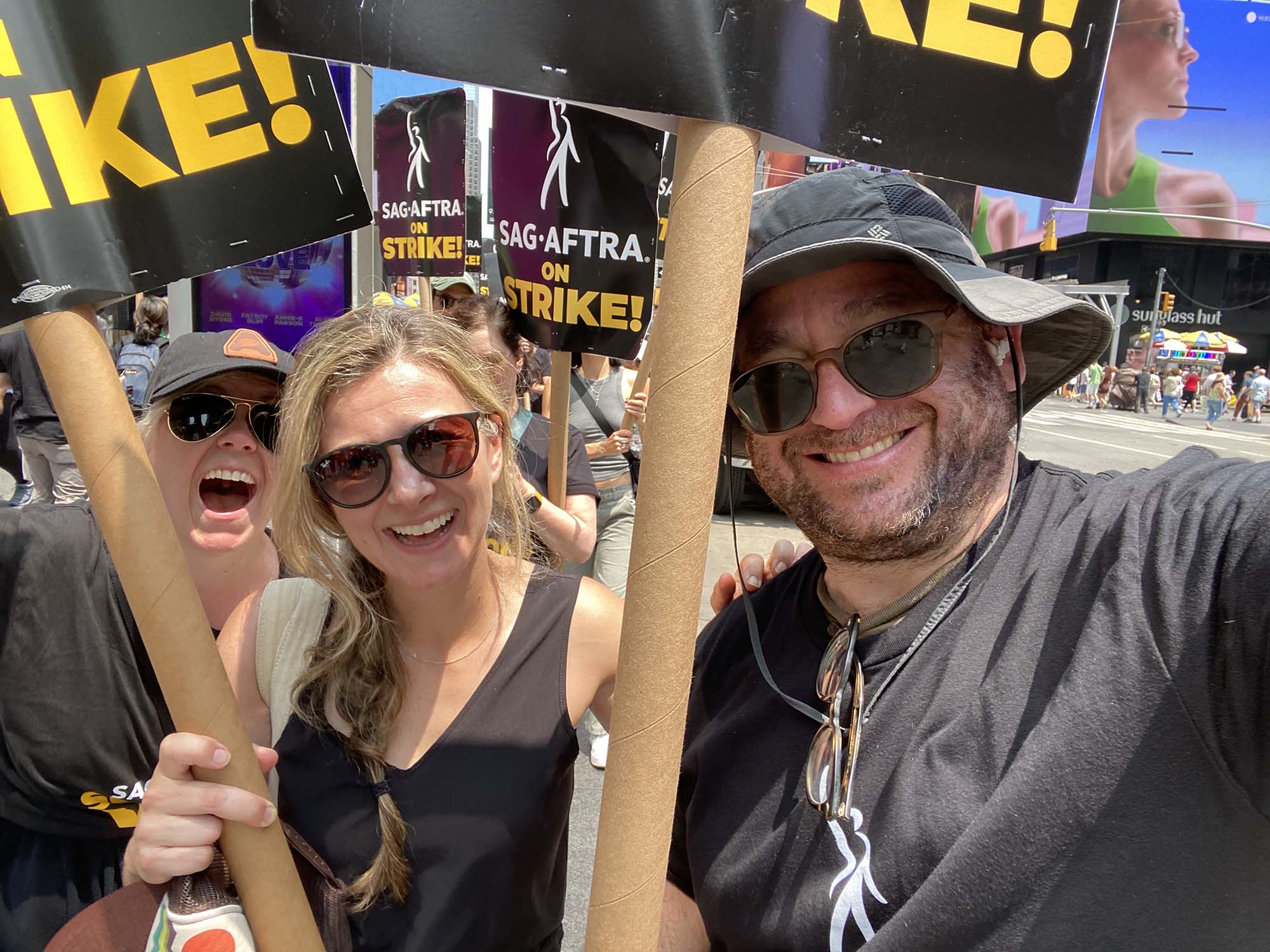 Photo: Dave Shalansky (right), is shown protesting during the strike. A white man holding a large sign and wearing a black cap, shirt, and sunglasses, poses with a few other people holding similar signs. A large group of protestors can be seen behind them holding similar signs.