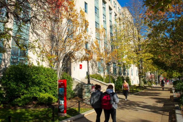 Photo: Stock photo of Boston. University's campus. Photo features students wearing falls sweatshirts and backpacks walking down the sidewalk in front of large campus buildings. The trees feature thinning yellow leaves.