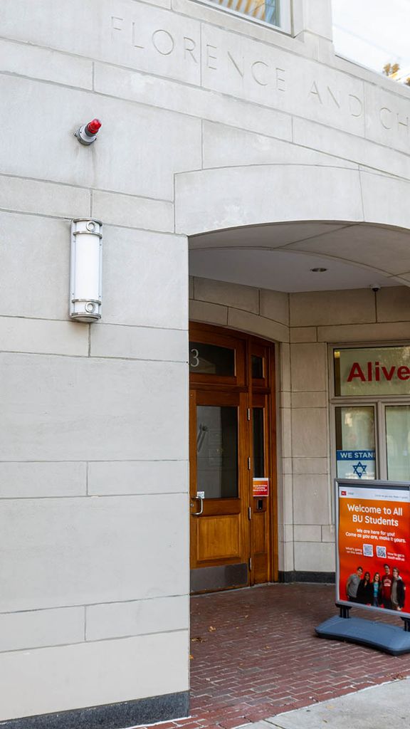 Photo: Exterior shot of BU's Hillel House. A grey brick building with wooden doors is shown. A red sign saying "Welcome to all BU Students" is placed in front of the door near the sidewalk and an Israeli flag can be seen in the left window of the building.
