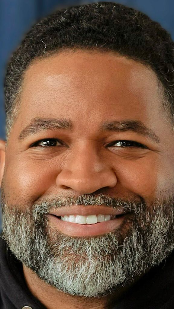 Photo: Jamil Smith, a man with a beard, smiles for the camera. He is wearing a black hoodie and is in front of a blue curtain background.