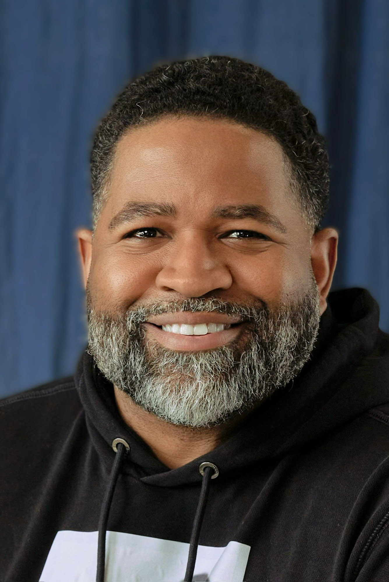 Photo: Jamil Smith, a man with a beard, smiles for the camera. He is wearing a black hoodie and is in front of a blue curtain background.