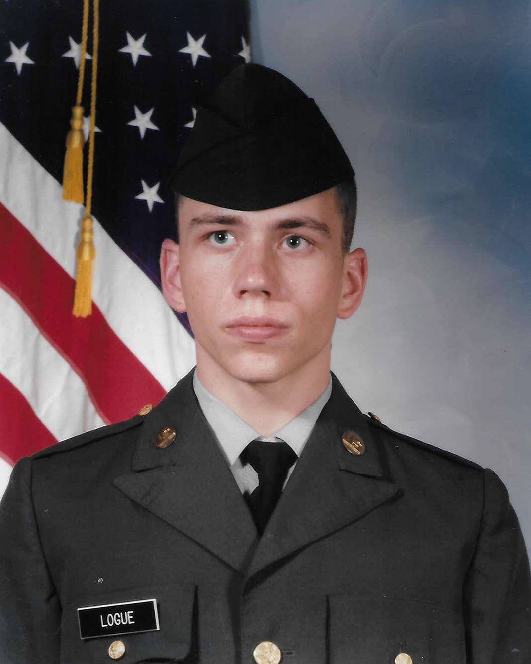 Photo: a young Logue is shown in a portrait. A young white man wearing a US Army reserve uniform stands in front of a large US flag behind him and looks stoic.